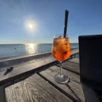 Ein Glas Aperol auf einem Holztisch vor dem Meer in der Sonne, daneben ein Laptop