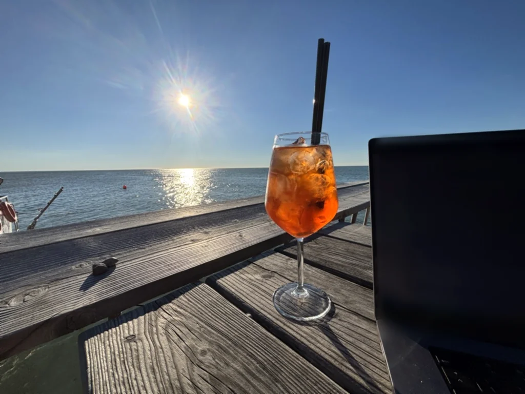Ein Glas Aperol auf einem Holztisch vor dem Meer in der Sonne, daneben ein Laptop