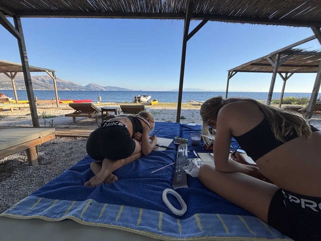 Zwei Mädchen beugen sich über ihre Aquarellblätter, im Hintergrund Kajaks und Meer