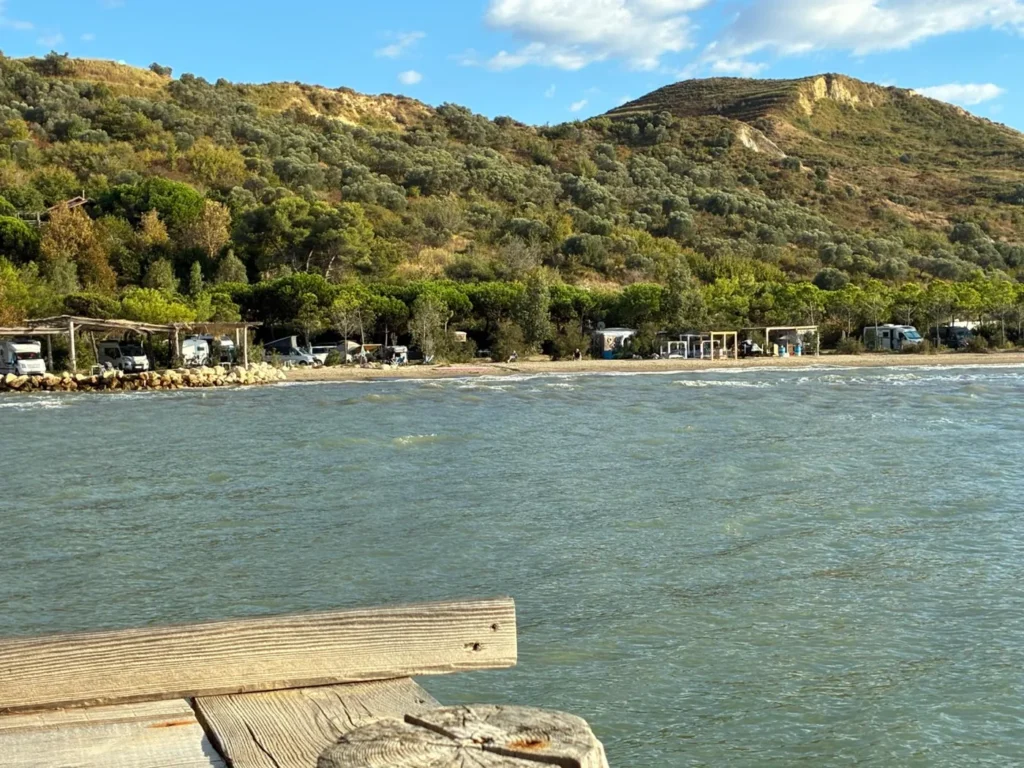 Blick vom Holzsteg über das Wasser auf den Campingplatz am Strand, dahinter ein grüner Hügel mit Olivenbäumen
