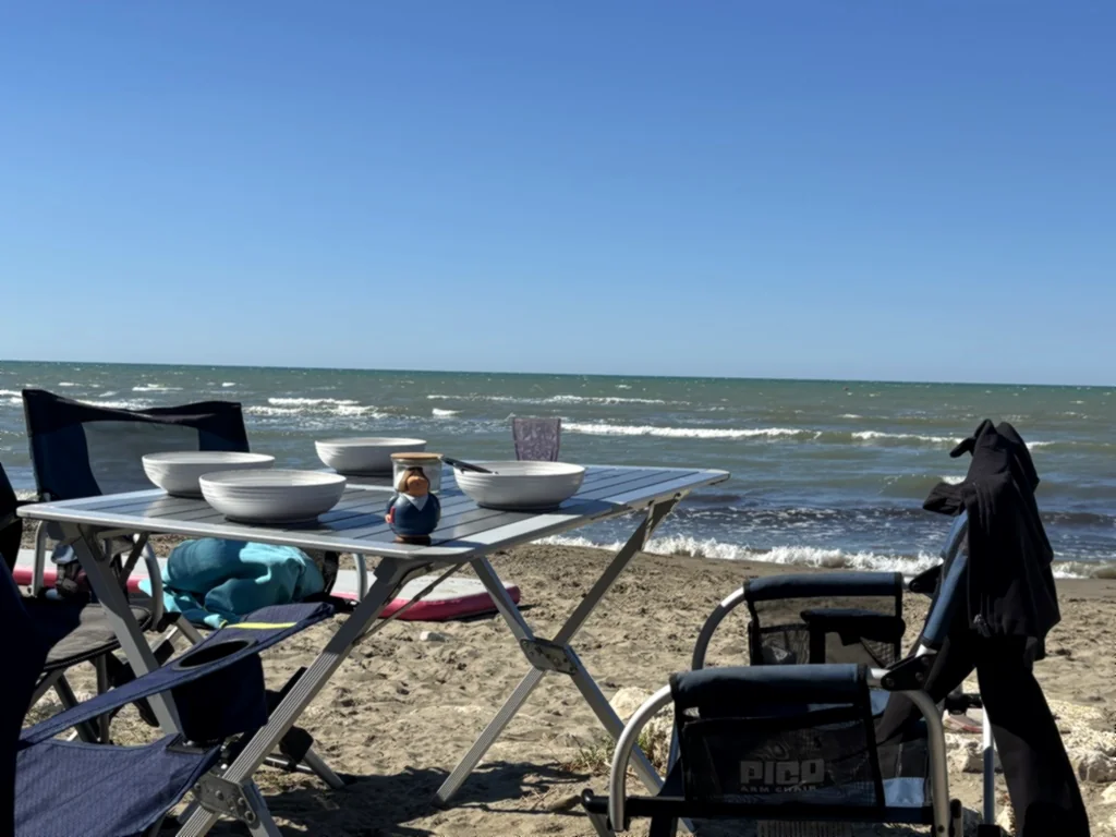 Campingtisch mit Pastatellern und Campingstühlen am Sandstrand, dahinter Wellen und blauer Himmel