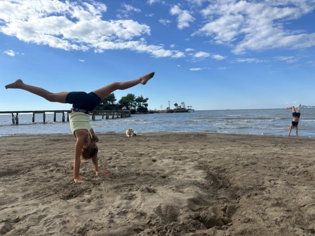 Zwei Kinder turnen am Strand, eines im Vordergrund, eines in der Ferne, ein Hund liegt dazwischen im Sand