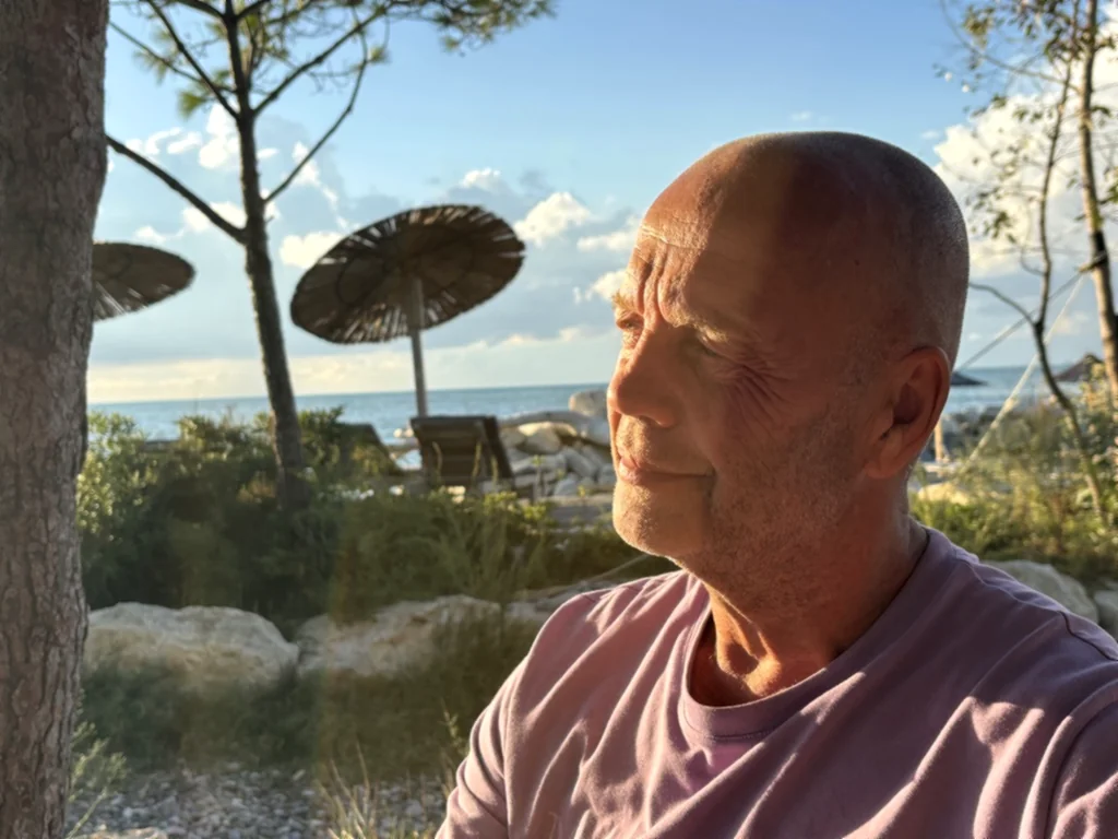 Heiner blickt im warmen Abendlicht aufs Meer, dahinter Strohschirme