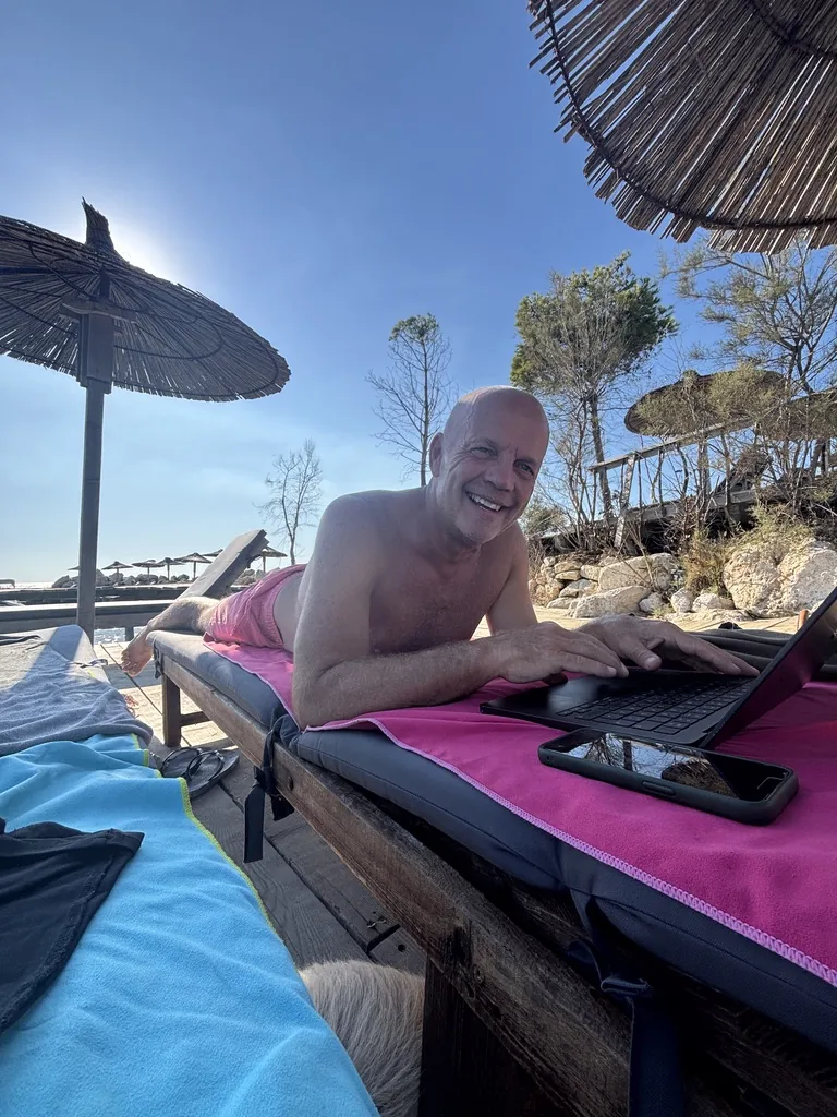 Heiner liegt lachend auf einer Liege mit pinkem Handtuch und tippt auf dem Laptop