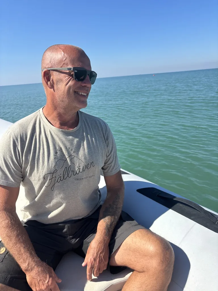 Heiner sitzt mit Sonnenbrille am Bootsrand und lächelt, dahinter das türkise Meer