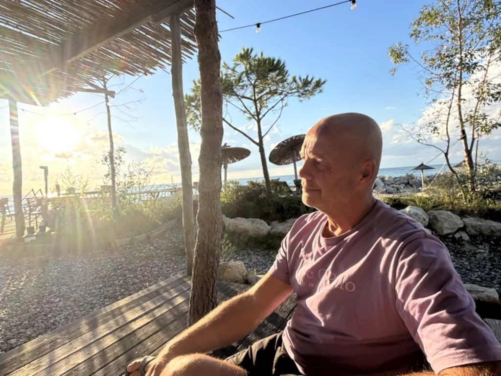 Heiner sitzt unter einem Schilfdach im Inselrestaurant, die tiefstehende Sonne strahlt ins Bild