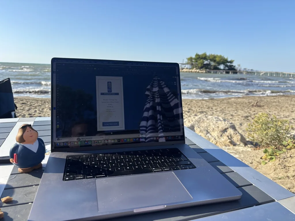 Aufgeklappter Laptop auf einem Tisch am Strand, dahinter Wellen und die kleine Insel mit Steg
