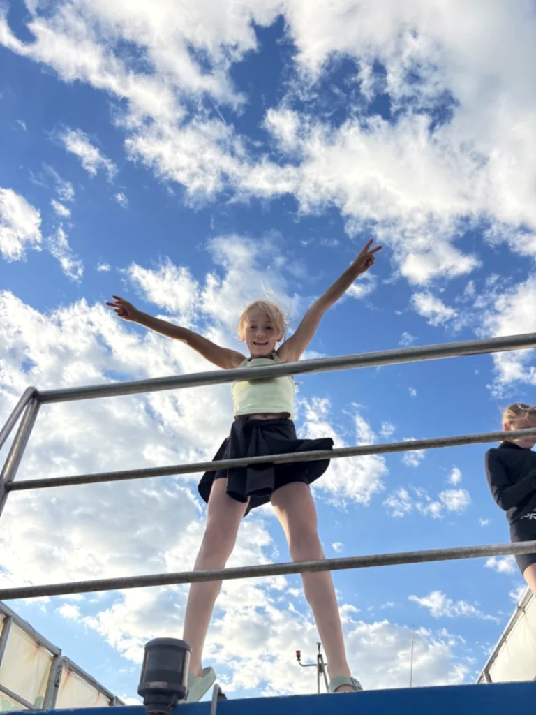 Ein Kind steht mit ausgestreckten Armen am Geländer auf dem Oberdeck des Bootes vor Wolkenhimmel
