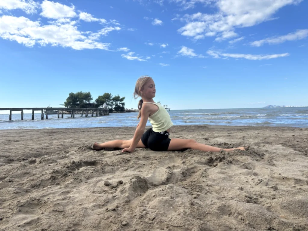 Ein lächelndes Kind sitzt im Spagat im Sand vor dem Meer