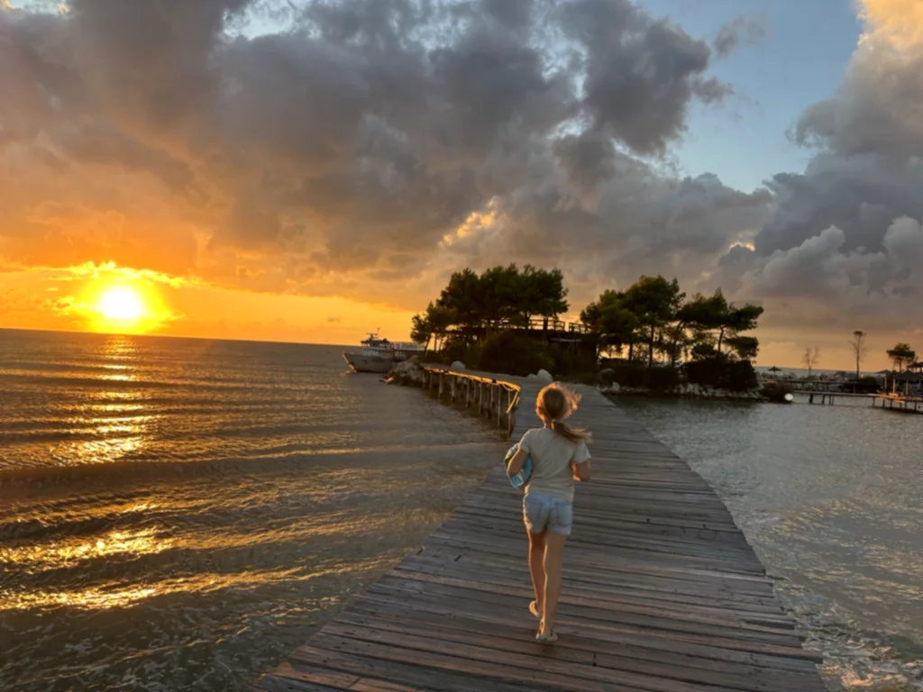 Ein Kind läuft über den Holzsteg zur Insel, links geht die Sonne orange über dem Meer unter