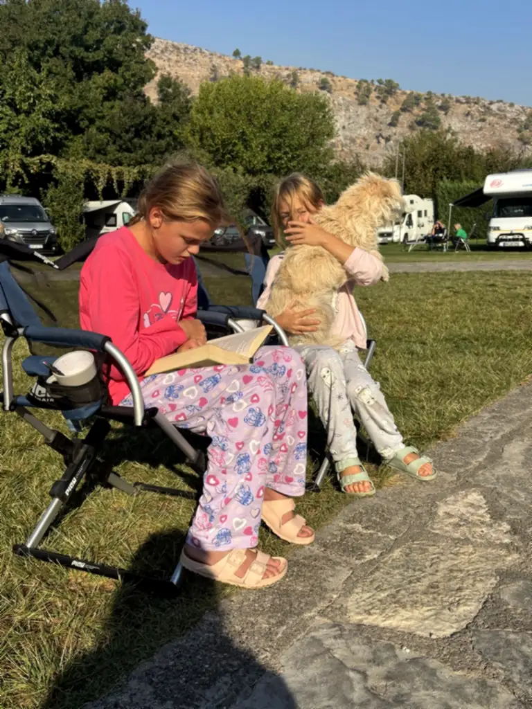 Zwei Mädchen im Pyjama sitzen im Campingstuhl, eine liest ein Buch, die andere kuschelt mit einem hellen Hund