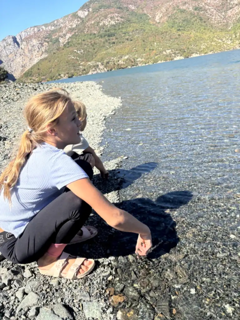 Zwei Kinder hocken am Kiesufer und greifen ins klare Wasser des Koman-Stausees