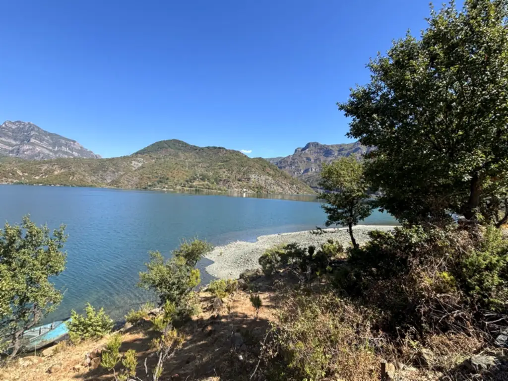 Kleine Kiesbucht am türkisblauen Koman-Stausee, umrahmt von Bäumen und Bergen