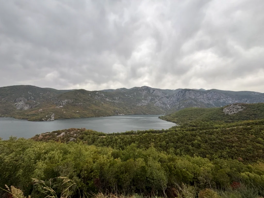 Grauer Koman-Stausee zwischen bewaldeten Bergen unter dichter Wolkendecke