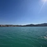 Türkises Meer mit Sonnenglitzern, dahinter eine felsige Küste unter blauem Himmel