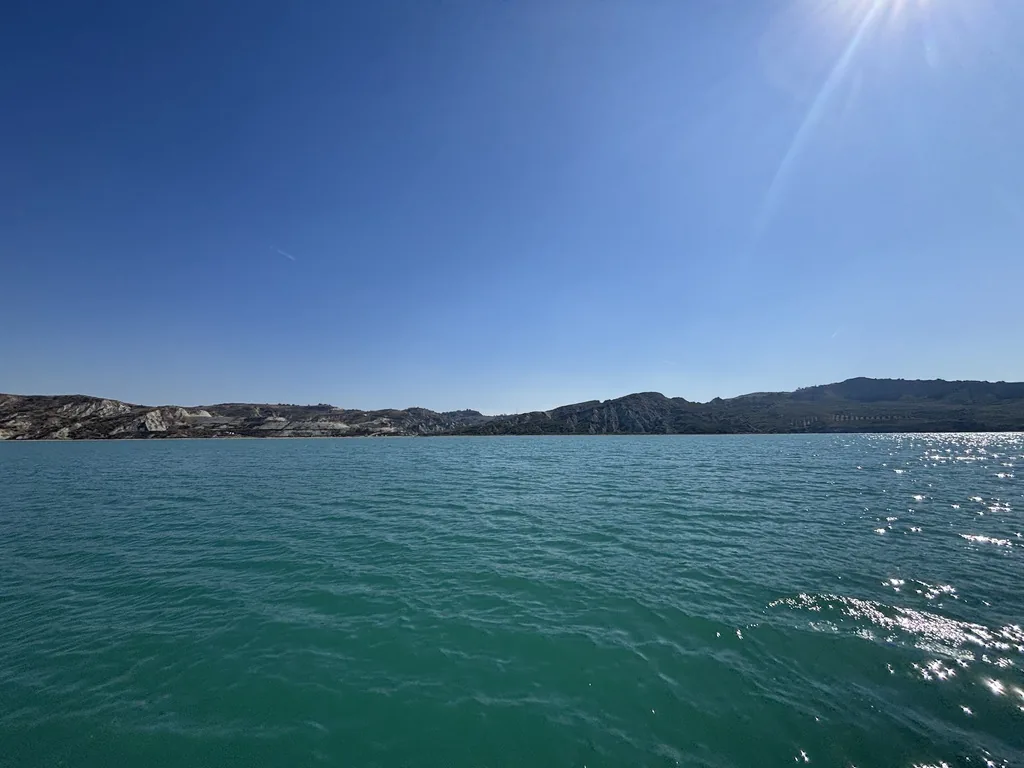 Türkises Meer mit Sonnenglitzern, dahinter eine felsige Küste unter blauem Himmel