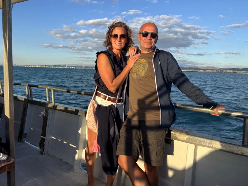 Lisa und Heiner stehen Arm in Arm an der Reling, dahinter Meer und Küste