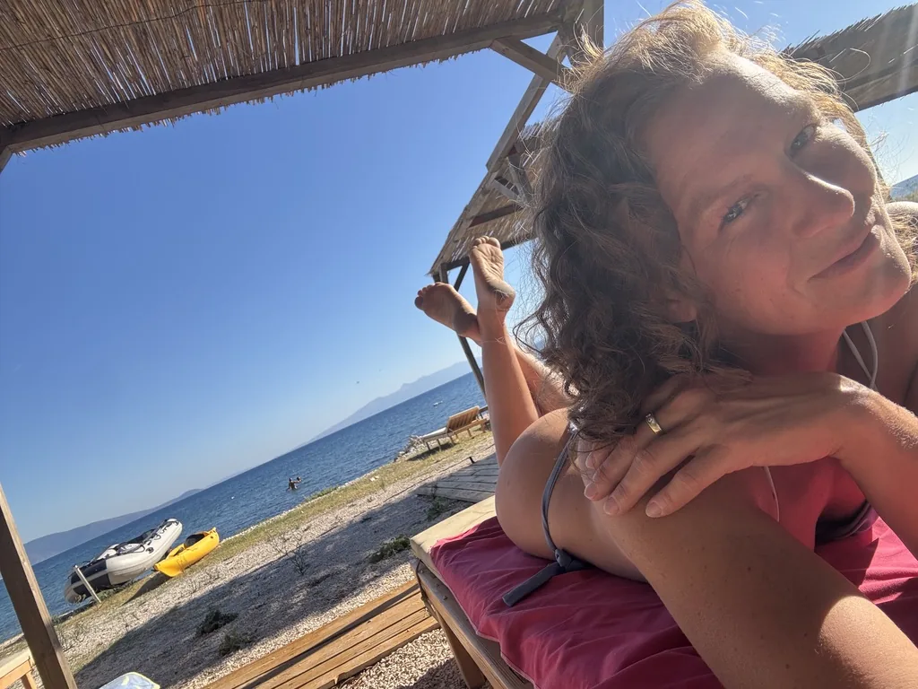 Lisa liegt lächelnd auf einer Liege unter dem Schilfdach, dahinter Kiesstrand und Meer