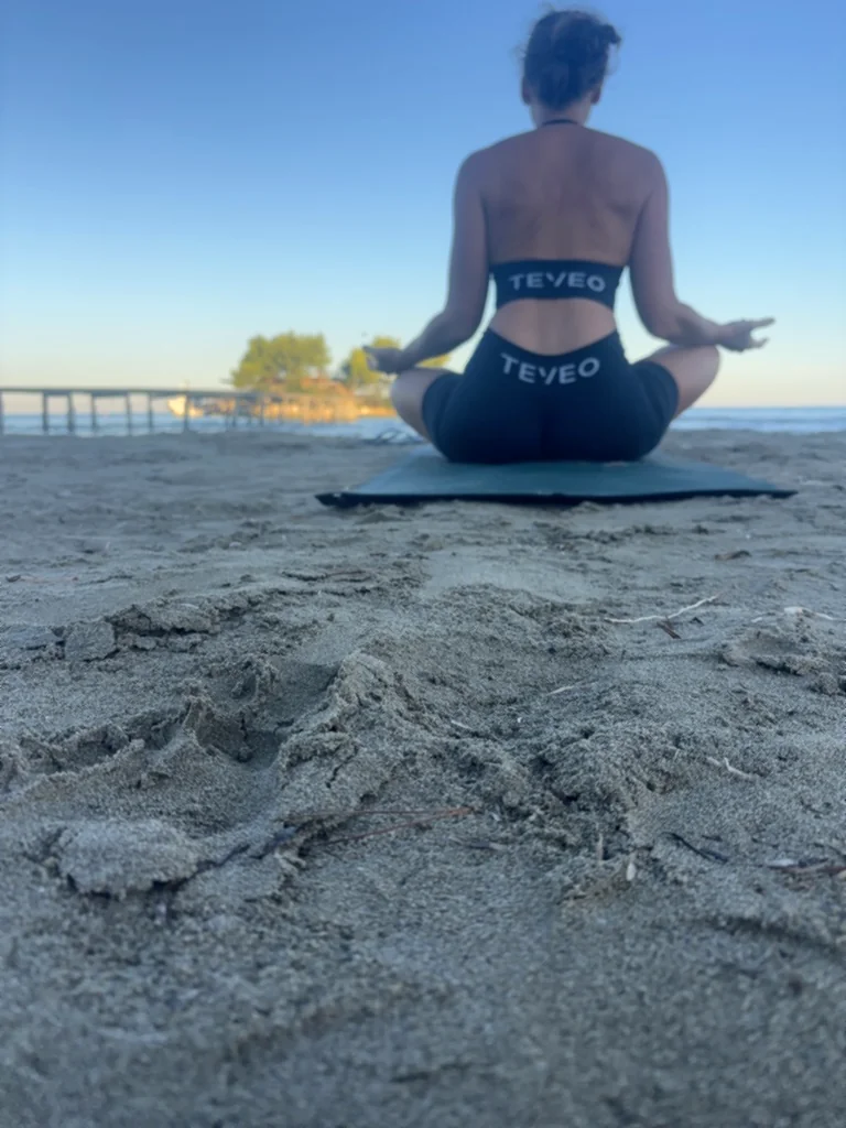 Lisa sitzt im Schneidersitz auf der Yogamatte im Sand, dahinter Steg und Insel im Morgenlicht