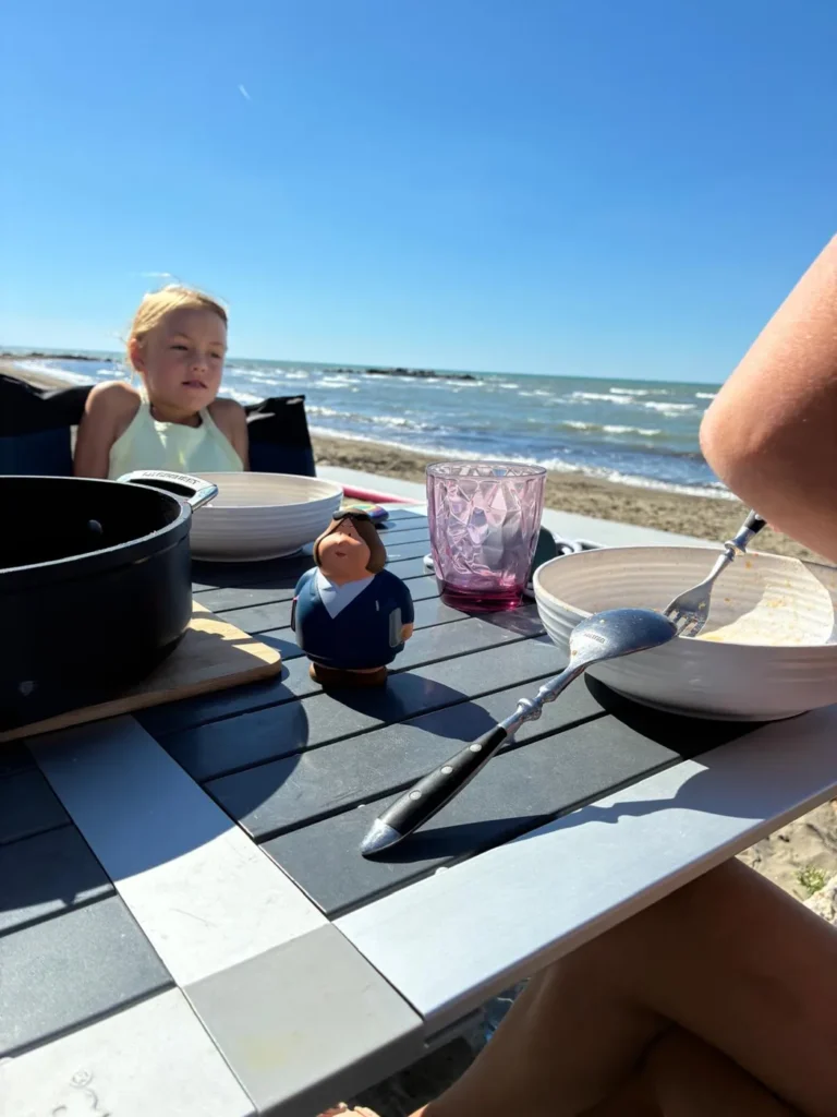 Gedeckter Campingtisch mit Topf und Schalen am Strand, im Hintergrund ein Mädchen und das Meer