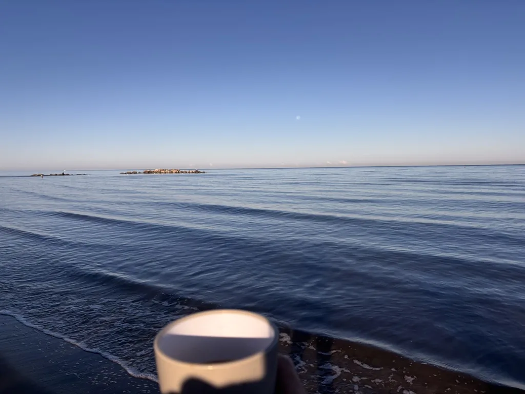Kaffeetasse im Vordergrund, dahinter das ruhige Meer im Morgenlicht mit dem Mond am Himmel