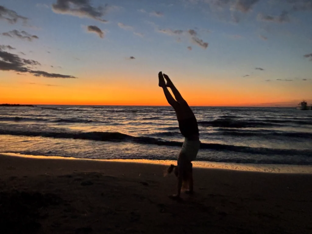 Silhouette eines Kindes, das am Strand in den Handstand schwingt, dahinter Wellen und Abendrot