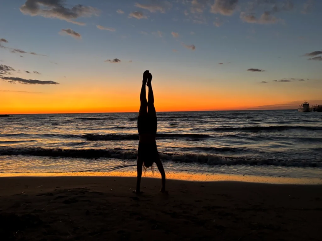 Silhouette eines Kindes im geraden Handstand am Wasser, rechts ein Boot am Horizont