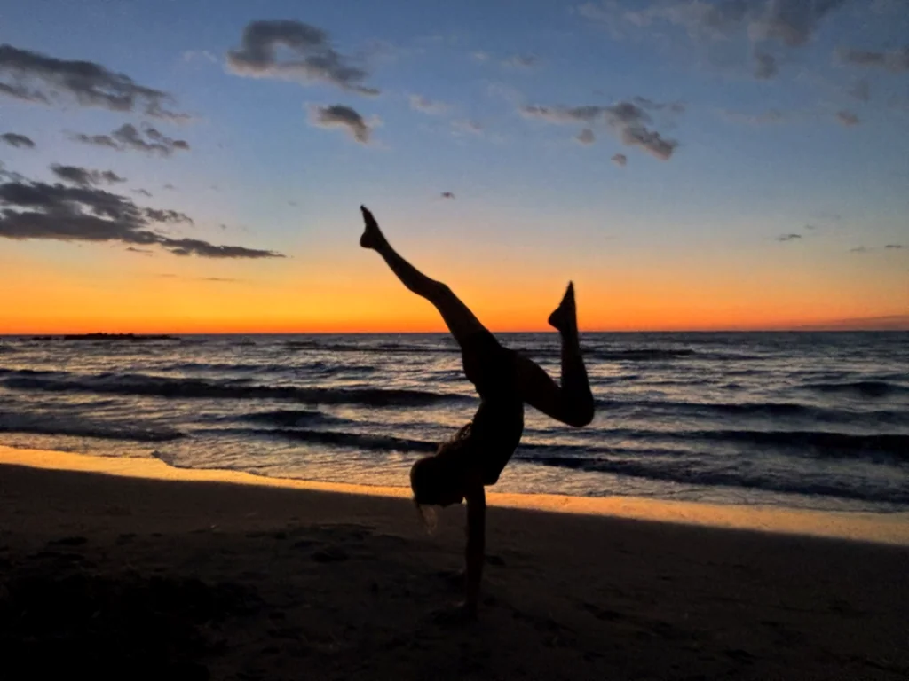 Silhouette eines Kindes im Handstand mit angewinkelten Beinen vor orangefarbenem Abendhimmel am Meer