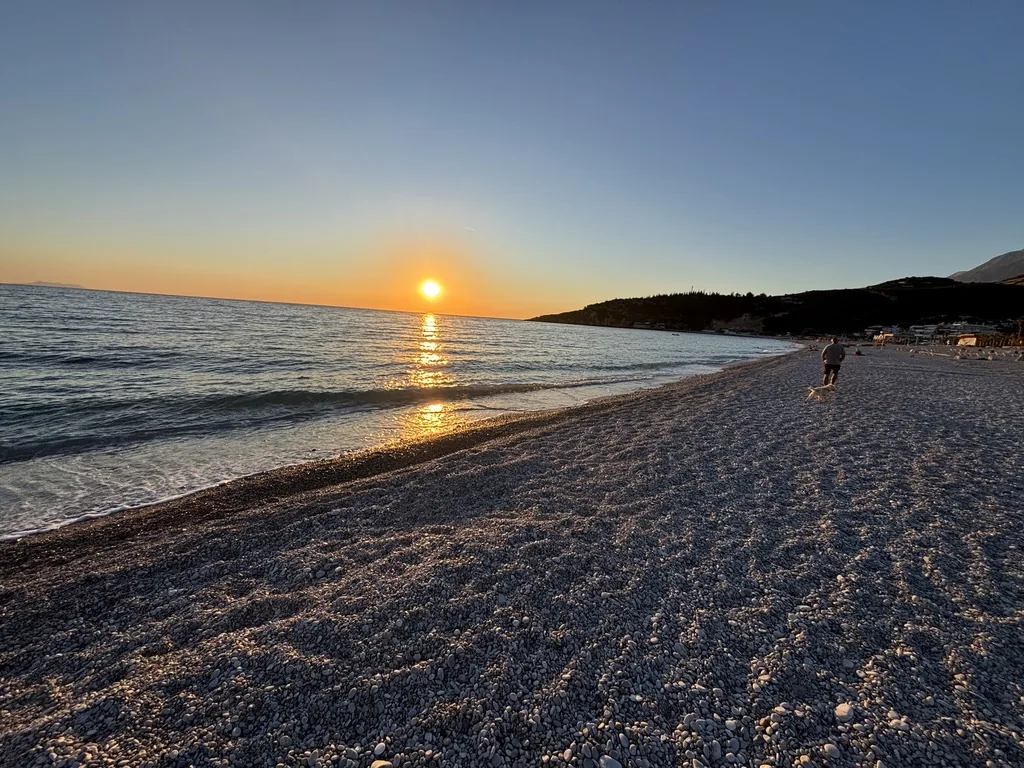 Die Sonne geht über dem Meer unter, am Kieselstrand läuft eine Person mit Hund