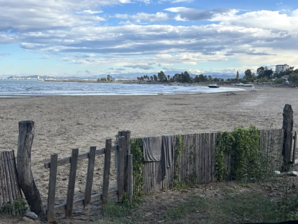 Sandstrand hinter einem Holzzaun mit Handtüchern, dahinter eine weite Bucht mit Booten und Bergen