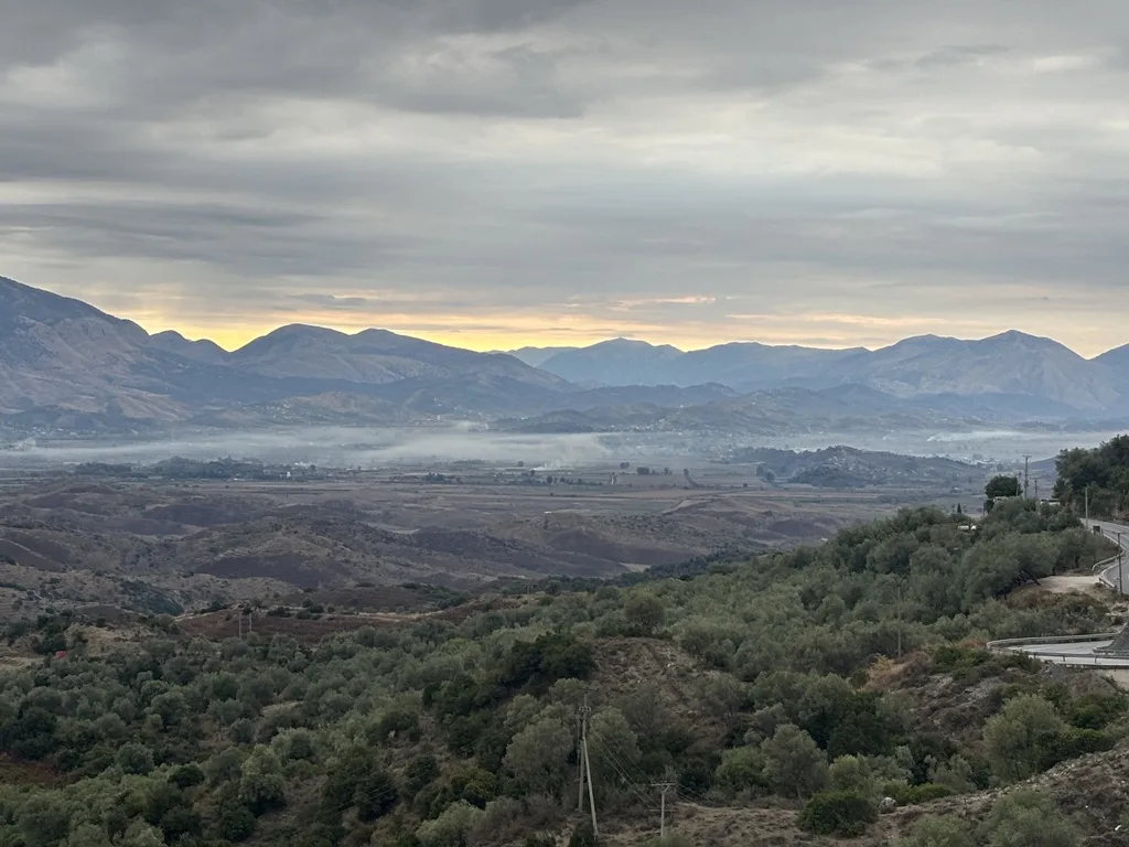 Nebelschwaden liegen über einer Ebene, dahinter Bergketten mit hellem Streifen am Horizont