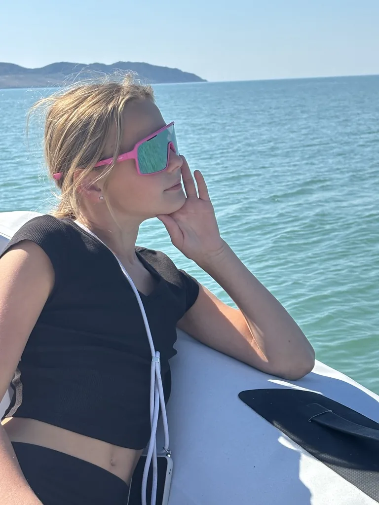 Anna-Luisa mit pinker Sportbrille lehnt am Bootsrand und blickt aufs Meer, hinten die Küste