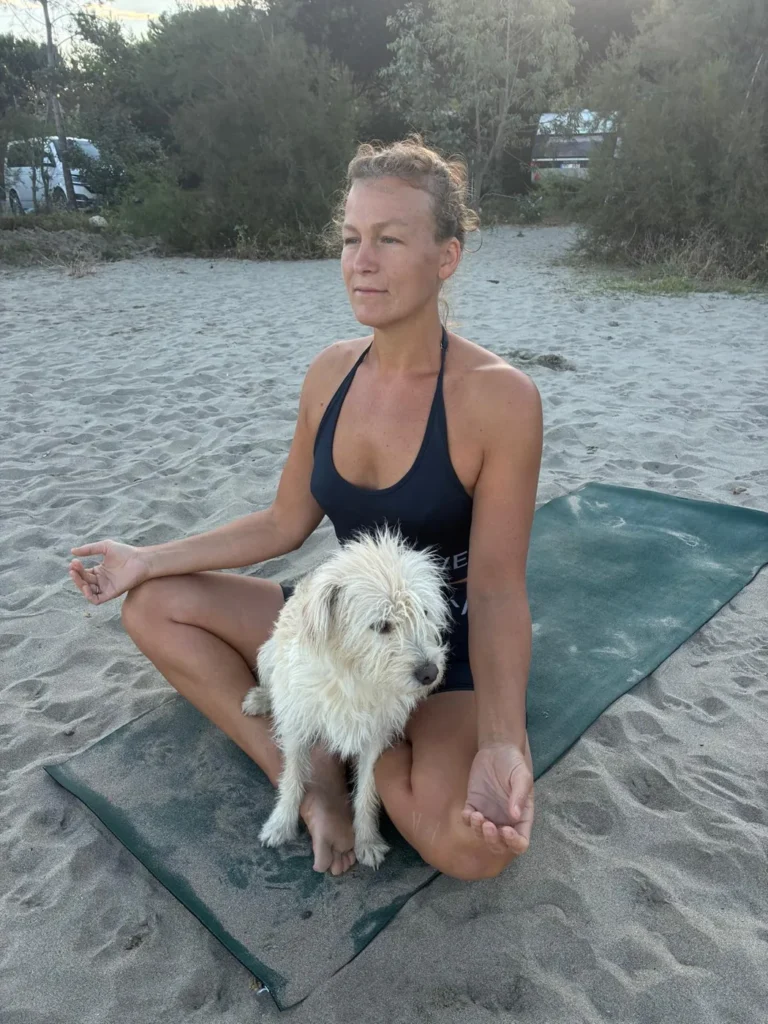 Lisa sitzt im Schneidersitz auf der Yogamatte am Strand, Chashew sitzt auf ihrem Schoß